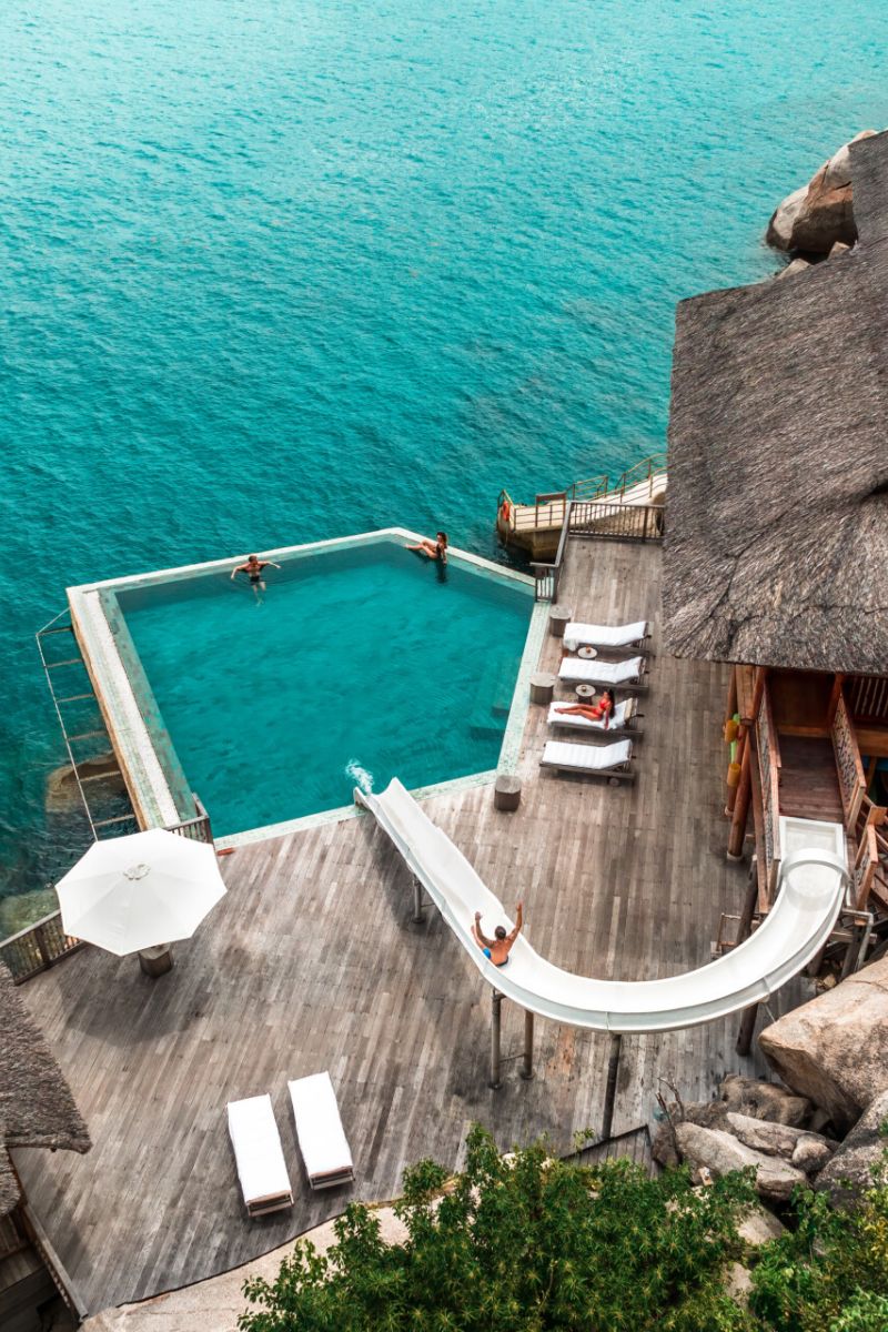 Aerial view of an oceanfront infinity pool with a water slide, sun loungers and a thatched-roof villa above turquoise sea